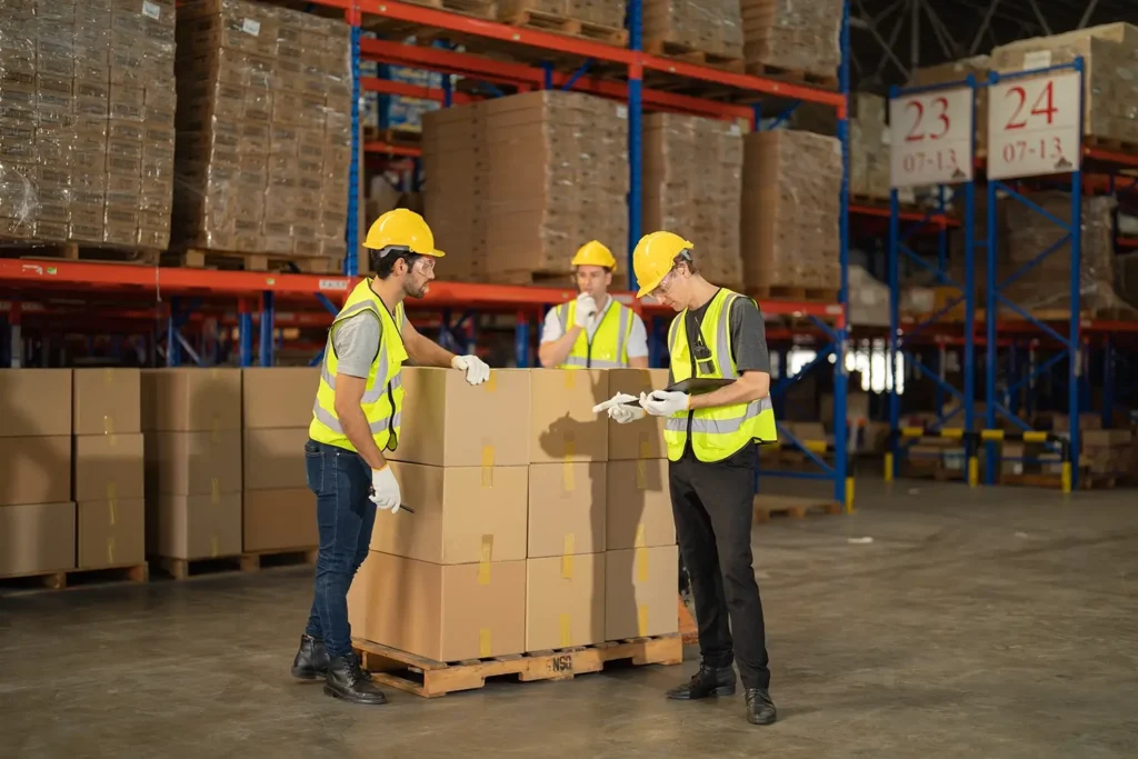 Drei Lagerarbeiter in Sicherheitswesten und mit Helmen kontrollieren Kisten auf einer Palette.